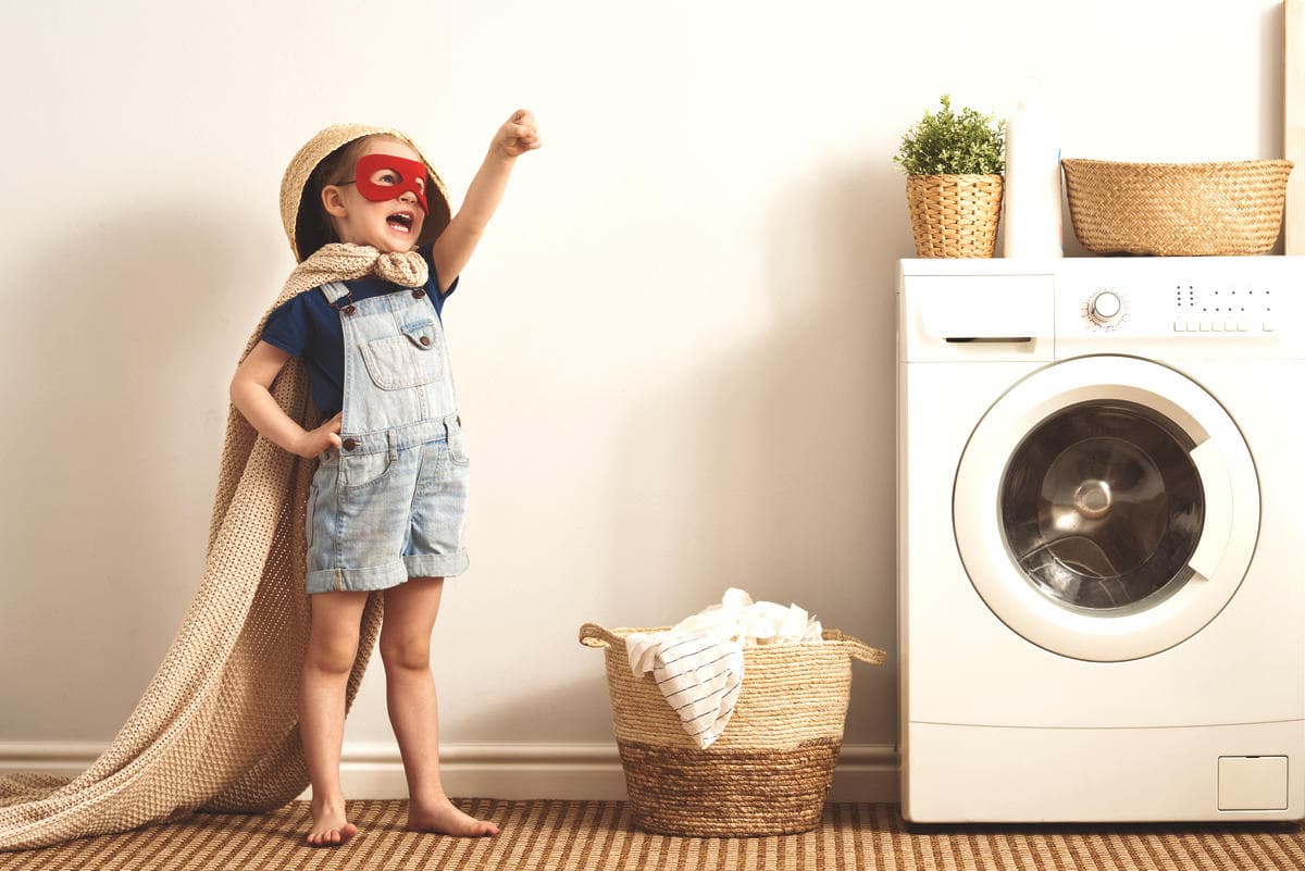 Child dressed as a superhero standing next to a washing machine and laundry basket, symbolizing a positive, practical approach to everyday home routines and learning through real life.