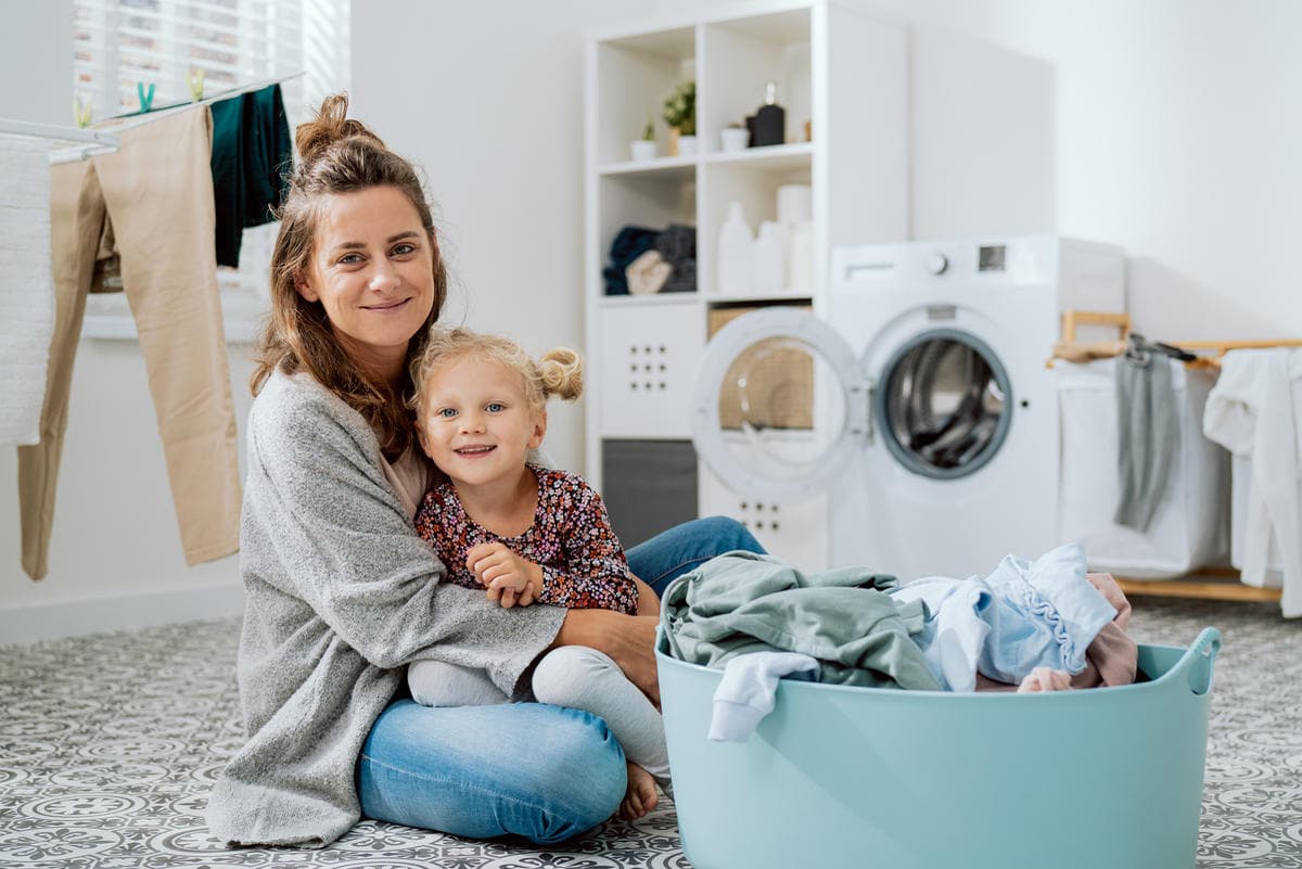 Parent and child sitting beside a laundry basket in a bright home laundry room, representing everyday cleaning routines and family-friendly home care.