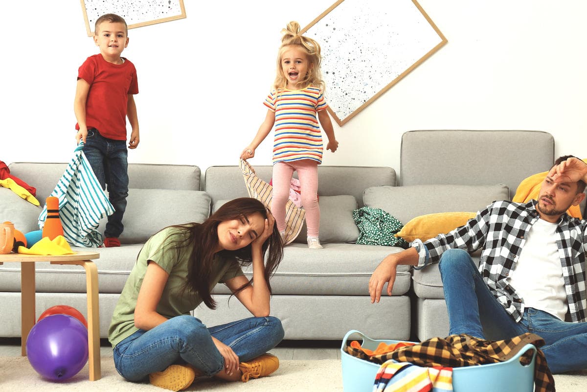Family in a lived-in living room with children playing and laundry visible, illustrating the reality of busy households and everyday home cleaning challenges.